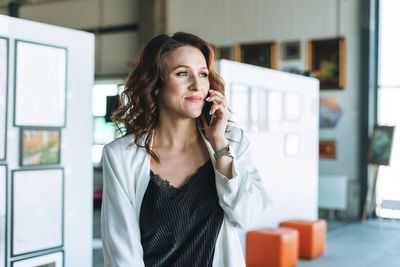 Charming young woman businesswoman designer artist in white suit using mobile phone in art gallery