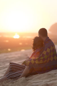 Thoughtful young couple wrapped in blanket while sitting at beach during sunset