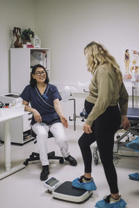 Full length of smiling female doctor looking at pregnant woman standing on weight scale in medical clinic