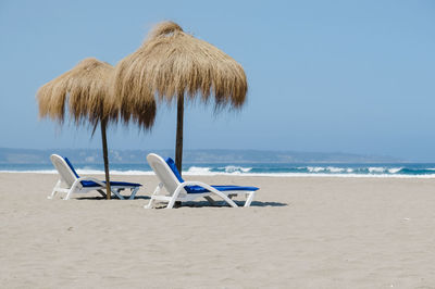 Deck chairs with thatched roof at beach against clear sky