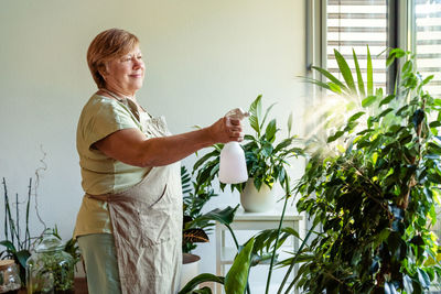 Adult senior retired woman taking care of plants at home spraying water at flowers. biophilia design