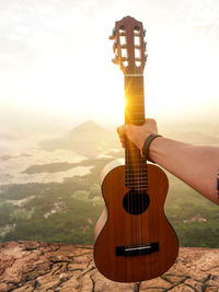 Cropped hand of man holding guitar on mountain against sky during sunset