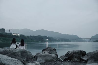 Rear view of female friends sitting on rock at lakeshore against sky