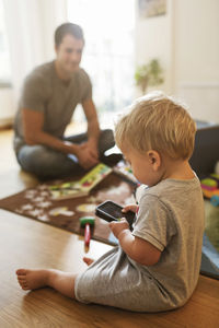 Baby boy using mobile phone on floor while father sitting with toys in background