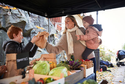 Smiling female carrying daughter and buying vegetables from market vendor