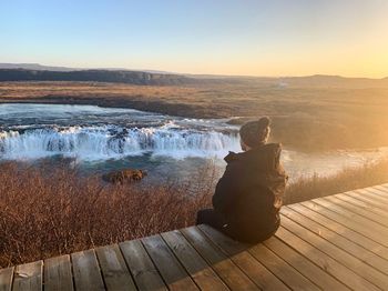 Rear view of man sitting on wood and looking at waterfall during sunset