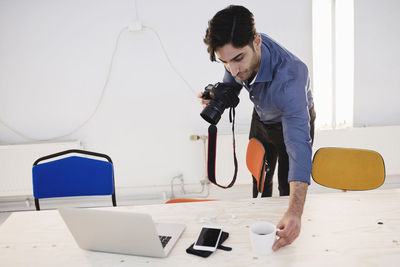 Male blogger placing coffee cup on desk while holding digital camera in office