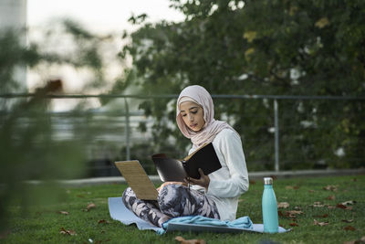 Full body of cheerful arab female in hijab with laptop on grassy lawn in park while consulting the agenda