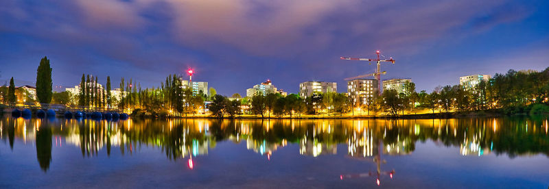 Illuminated buildings by lake against sky in city