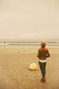 Full length rear view of woman walking by buoys at beach against sky