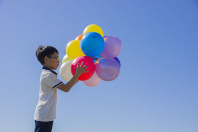 Boy holding multi colored balloons while standing against clear blue sky