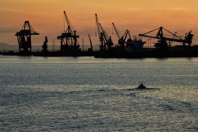 Silhouette cranes at harbor against sky during sunset