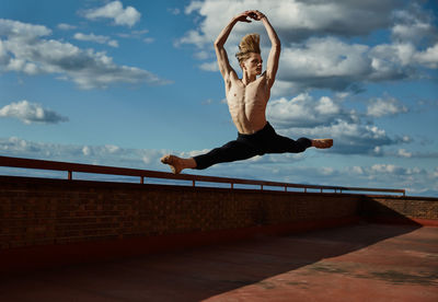 Masterful young blond ballet dancer in black leotard performing exciting jump with raising hands up on roof of building on sky background in sunny day