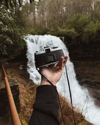 Cropped hand of person holding camera against waterfall