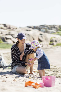 Baby girl pouring sand in mother's hand at beach on sunny day