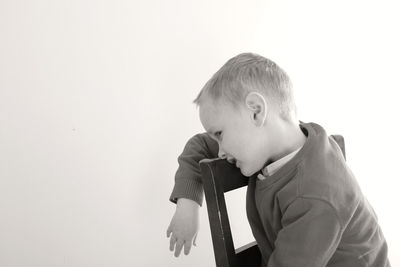 Boy leaning while sitting on chair against wall