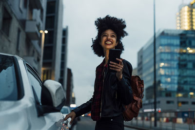 Cheerful young woman with mobile phone standing by taxi at dusk