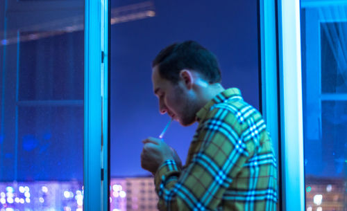 Man smoking cigarette while standing by window at dusk