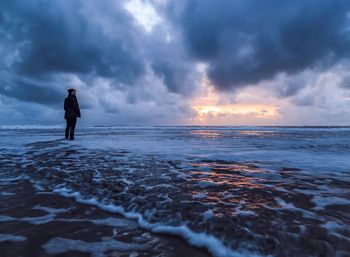 Rear view of person standing in the surf against cloudy sky