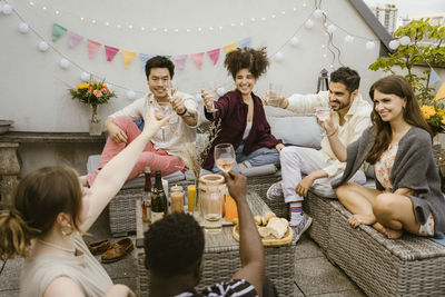 Group of multiracial friends toasting drinks while sitting together in balcony