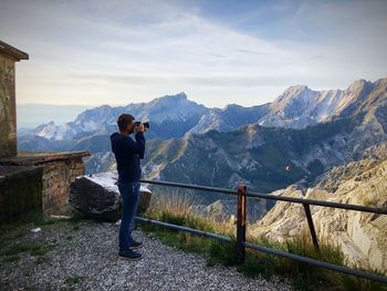 Man photographing while standing by railing against mountain range 