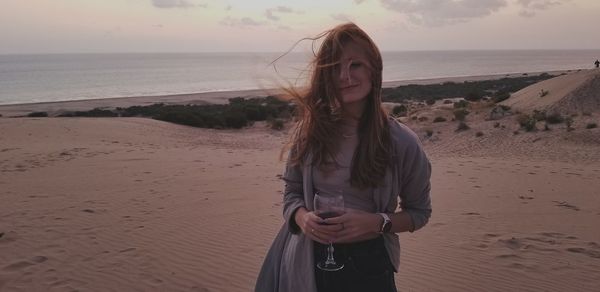 Woman having red wine while standing at beach during sunset