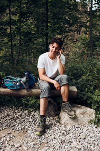 Young man with backpack talking on phone having break sitting on trunk during hike in a mountains