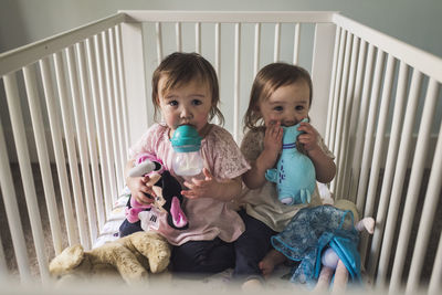 Portrait of twin toddler girls drinking milk in bedroom crib