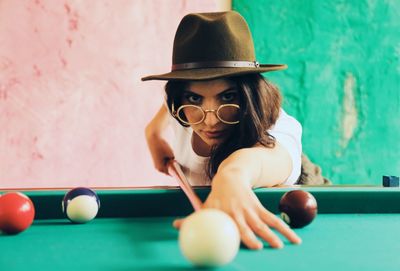 A woman wearing sexy hat and round glasses playing pool to a colorful pastel background