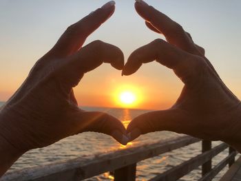 Cropped hands making heart shape against sea during sunset