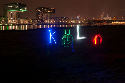 Illuminated text on riverbank against modern buildings at night