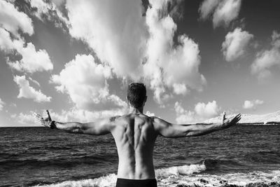 Rear view of shirtless man with arms outstretched standing by sea