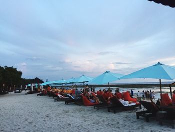 People relaxing under parasols at beach against sky during sunset