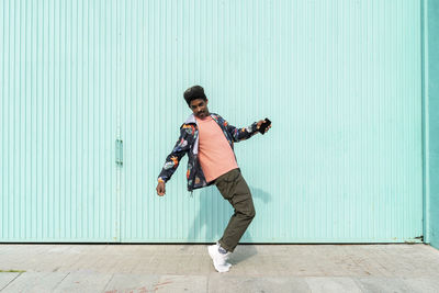 Ecstatic man holding smart phone while dancing in front of turquoise wall