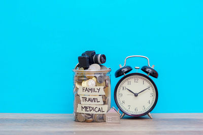 Close-up of alarm clock and coins in jar on table against blue background