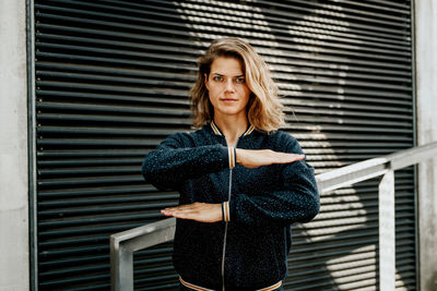 Portrait of woman gesturing equal sign while standing against closed shutter