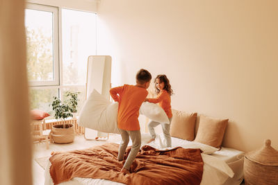 Funny happy kids brother and sister in pajamas playing fighting with pillows in cozy bedroom at home