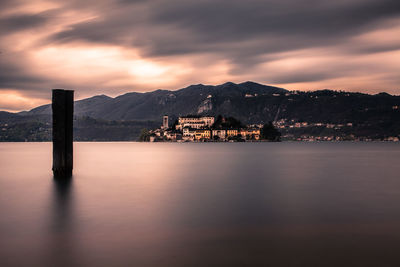 Wooden post in river by mountains against cloudy sky during sunset