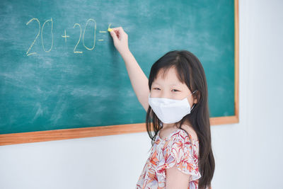 Little asian schoolgirl wearing fcial mask writing answer on blackboard in classroom at school.