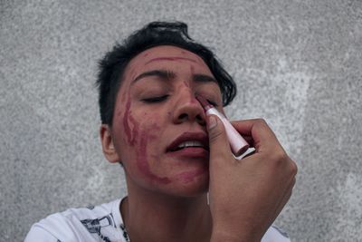 Close-up of young woman applying lipstick on face against wall