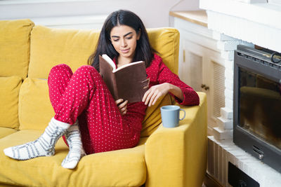 Young woman sitting on sofa reading book at home