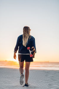 Rear view of young woman with skateboard walking at beach against clear sky during sunset