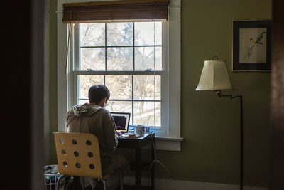 A man sits at desk in front of window in bedroom working on a computer