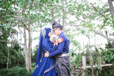 Side view of bride and groom embracing while standing against trees