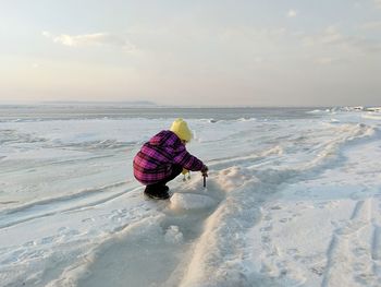 Rear view of girl playing with ice by sea against sky during winter