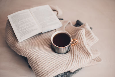 Cup of coffee stay on stack of knitted textile sweater in bed with paper open book. winter season.