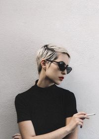 Close-up of woman wearing sunglasses holding cigarette standing by wall outdoors