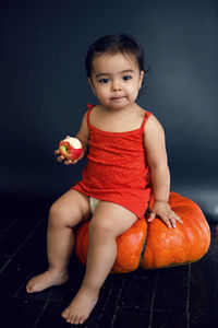 Baby girl in red tank top eating ripe red apple on black background in studio