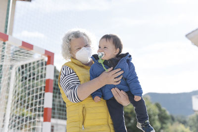 Grandmother wearing a medical mask with little grandson in her arms in a park on sunny day