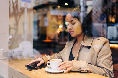 Young woman using phone while holding coffee at cafe